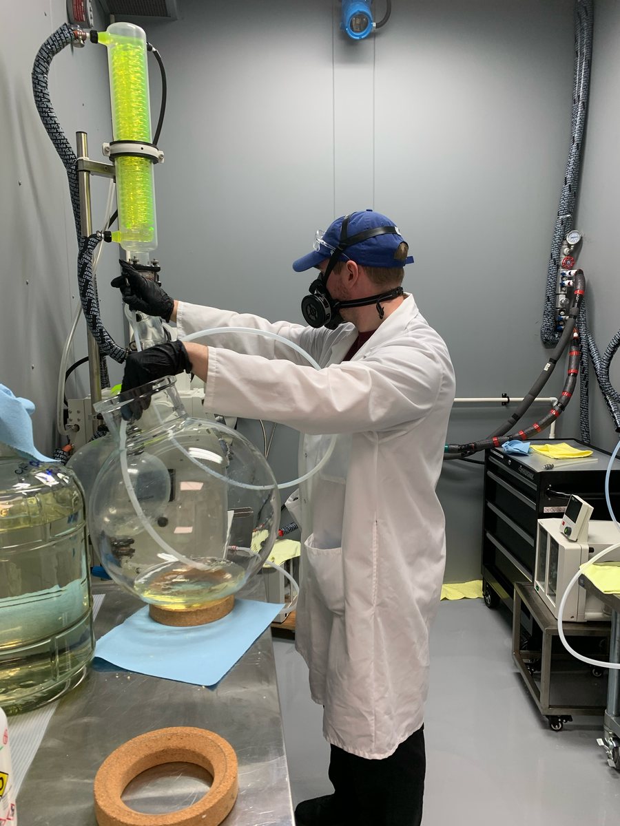 A technician in a lab coat, respirator, safety glasses and nitrile gloves lowering a large round-bottom flask on a rotary evaporator
